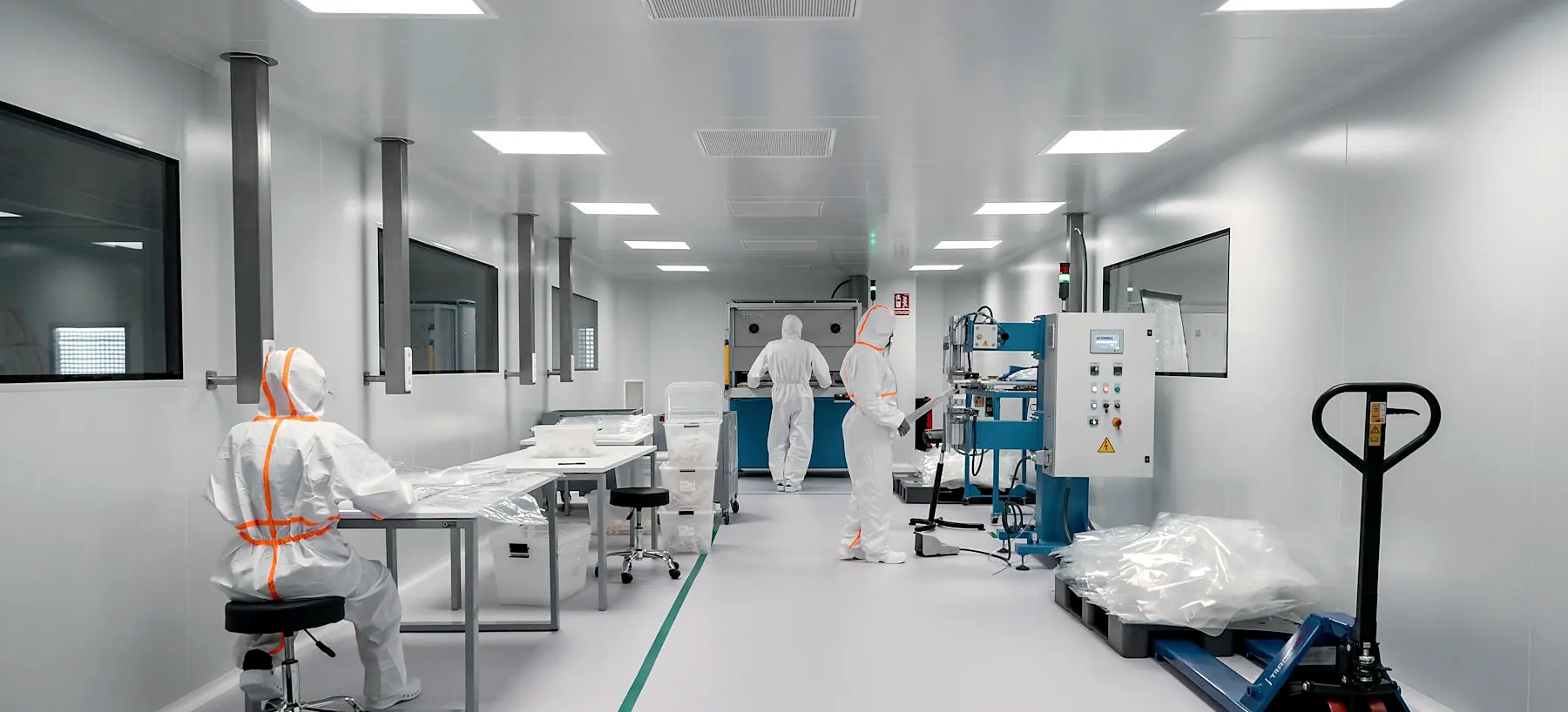 A brightly lit cleanroom with stainless benches and workers in white coveralls