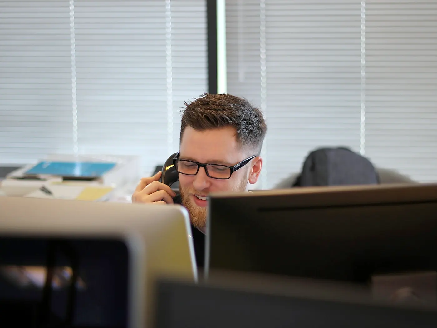 A man in glasses speaking on a desk handset behind his monitors
